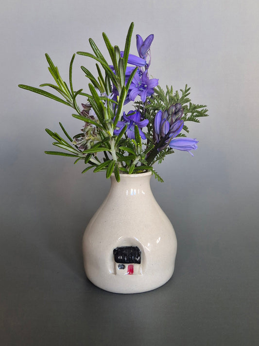 Small white ceramic bottle or bud vase decorated with a small house and filled with wildflowers. On a plain background
