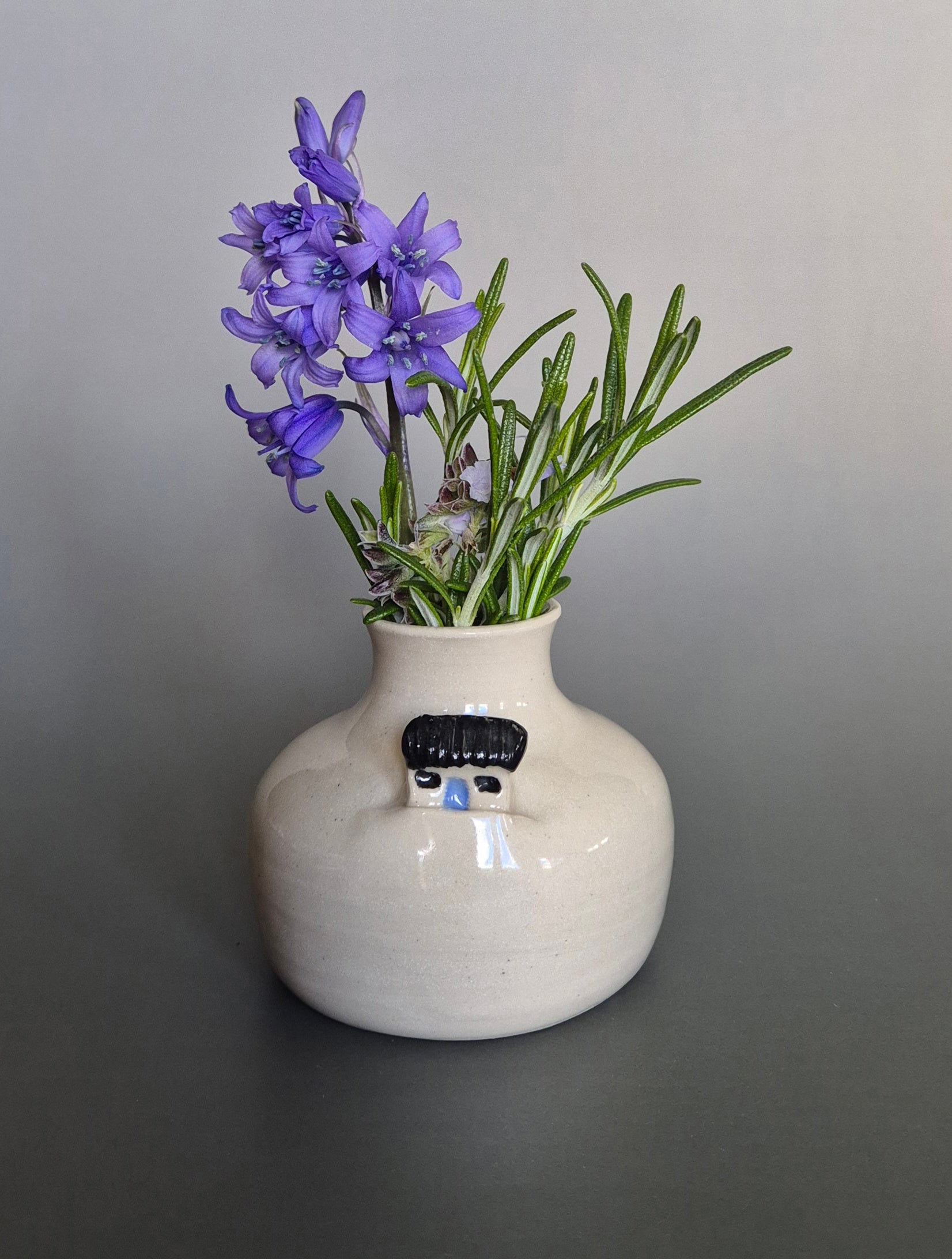 Small white ceramic bottle or bud vase decorated with a small house and filled with wildflowers. On a plain background