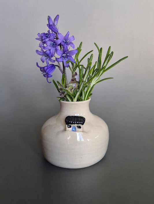 Small white ceramic bottle or bud vase decorated with a small house and filled with wildflowers. On a plain background