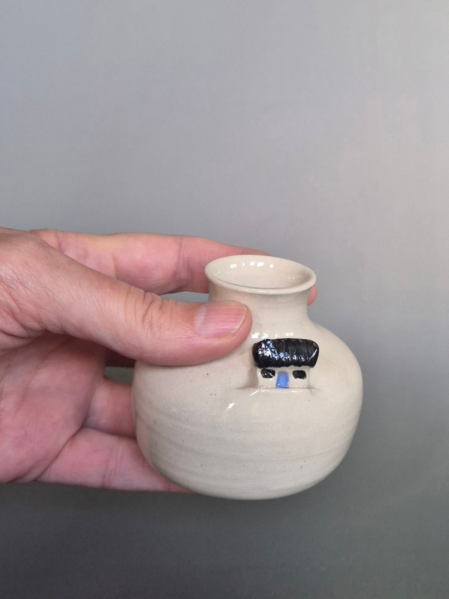 A hand holding a small white ceramic bottle or bud vase. On a plain background