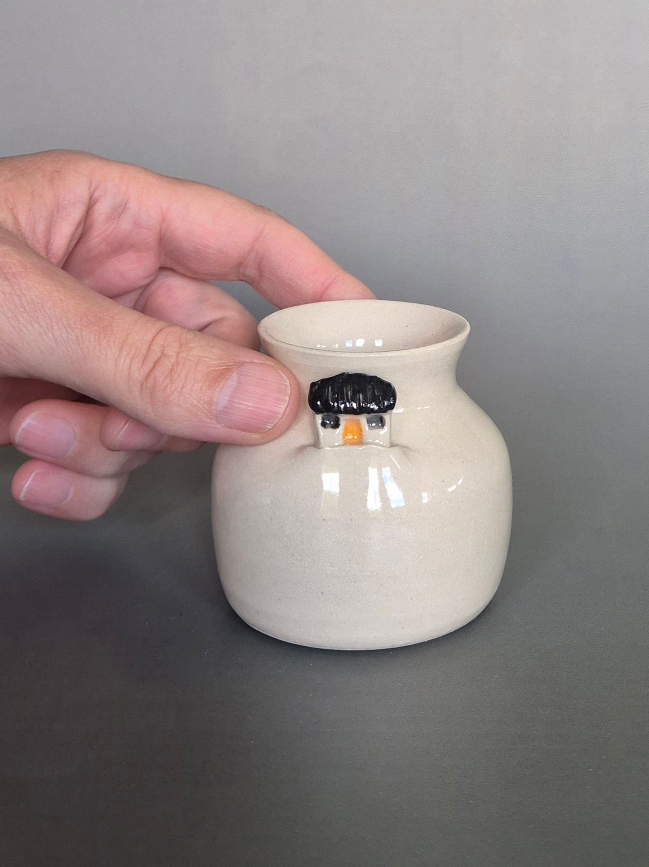 A hand holding a small white ceramic bottle or bud vase. On a plain background