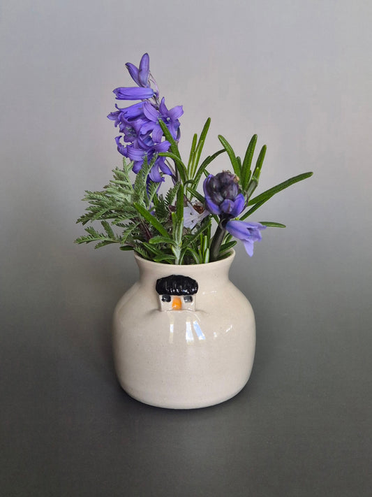 Small white ceramic bottle or bud vase decorated with a small house and filled with wildflowers. On a plain background