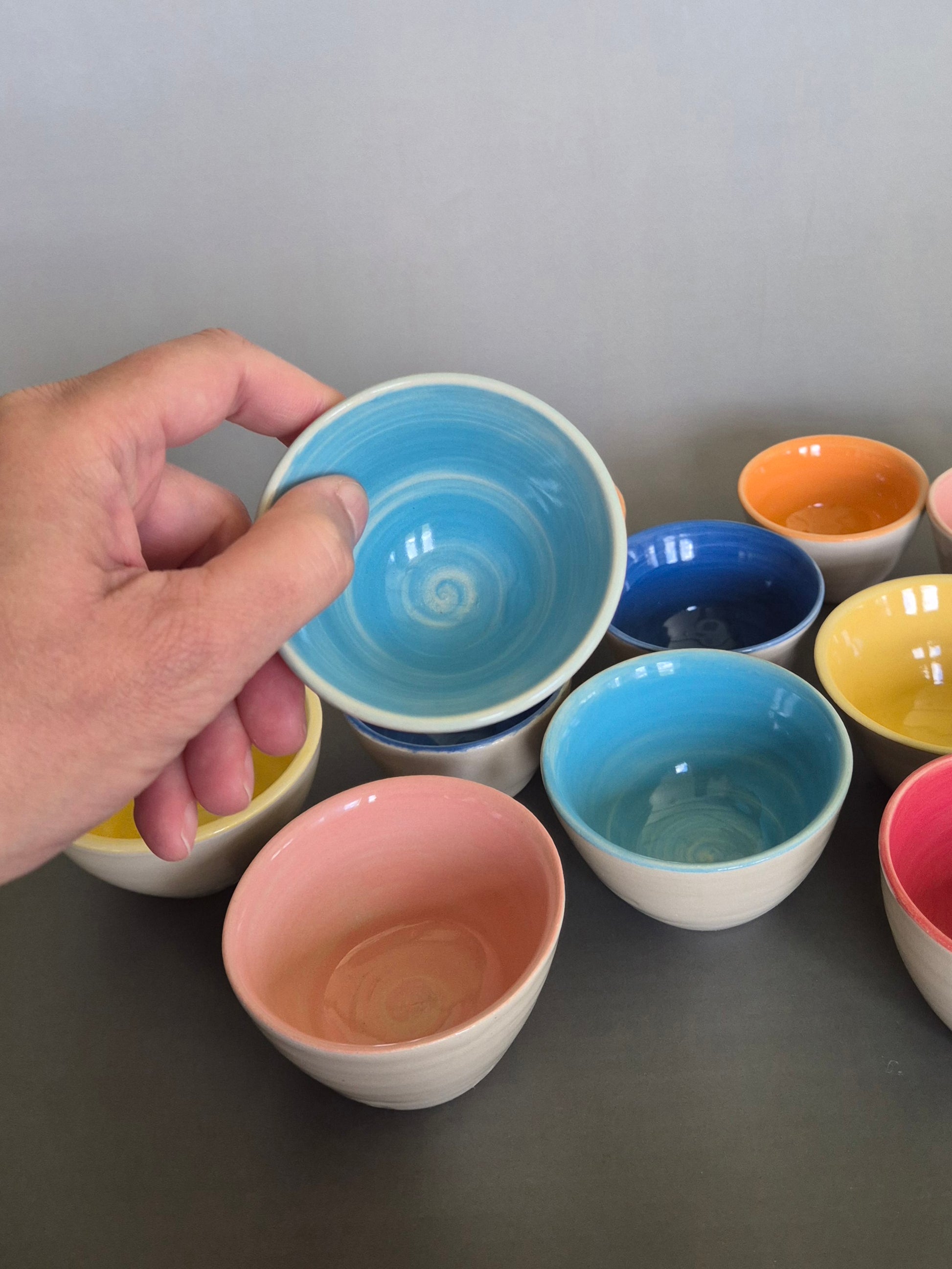 Hand holding a blue ceramic bowl among other colorful bowls on a gray surface.