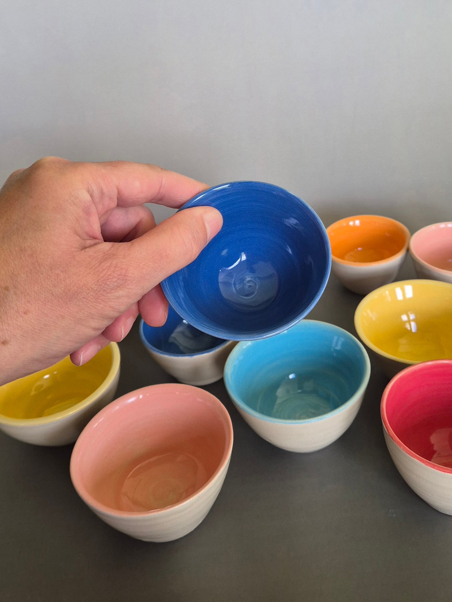 A person holding a brightly coloured condiment bowl with other colourful bowls in the background. On a plain background.
