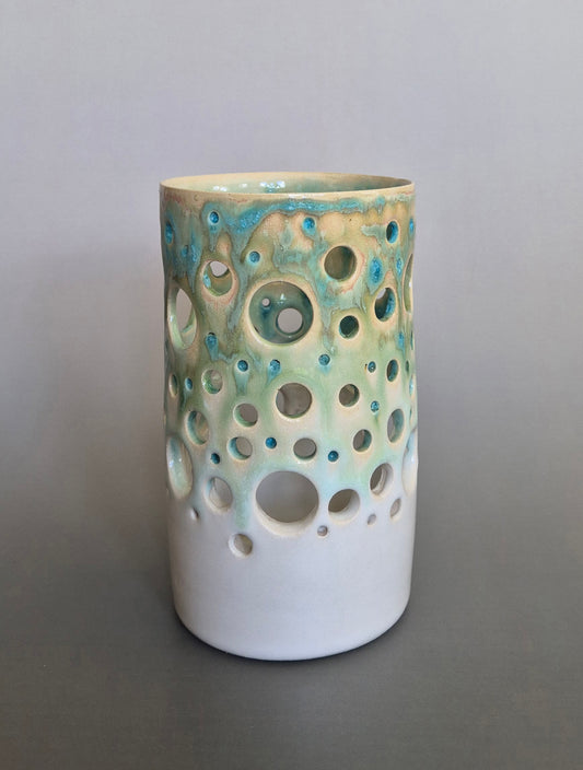 Tall ceramic cylinder pierced with holes and glazed with a flowing blue/green glaze. On a plain background
