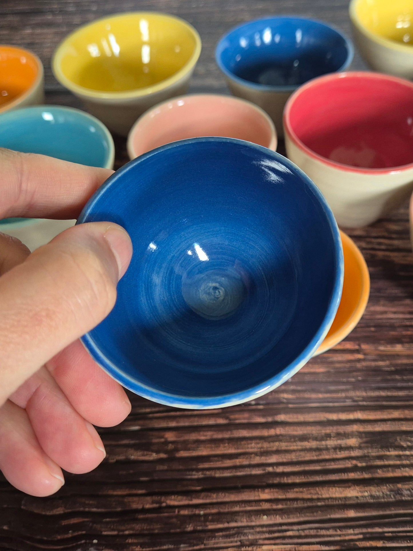 A person holding a brightly coloured condiment bowl with other colourful bowls in the background. On a wood table.