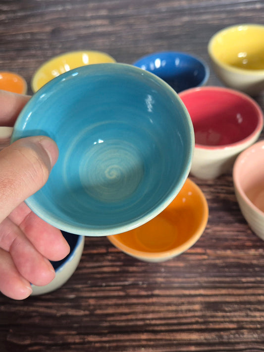 A person holding a brightly coloured condiment bowl with other colourful bowls in the background. On a wood table.

