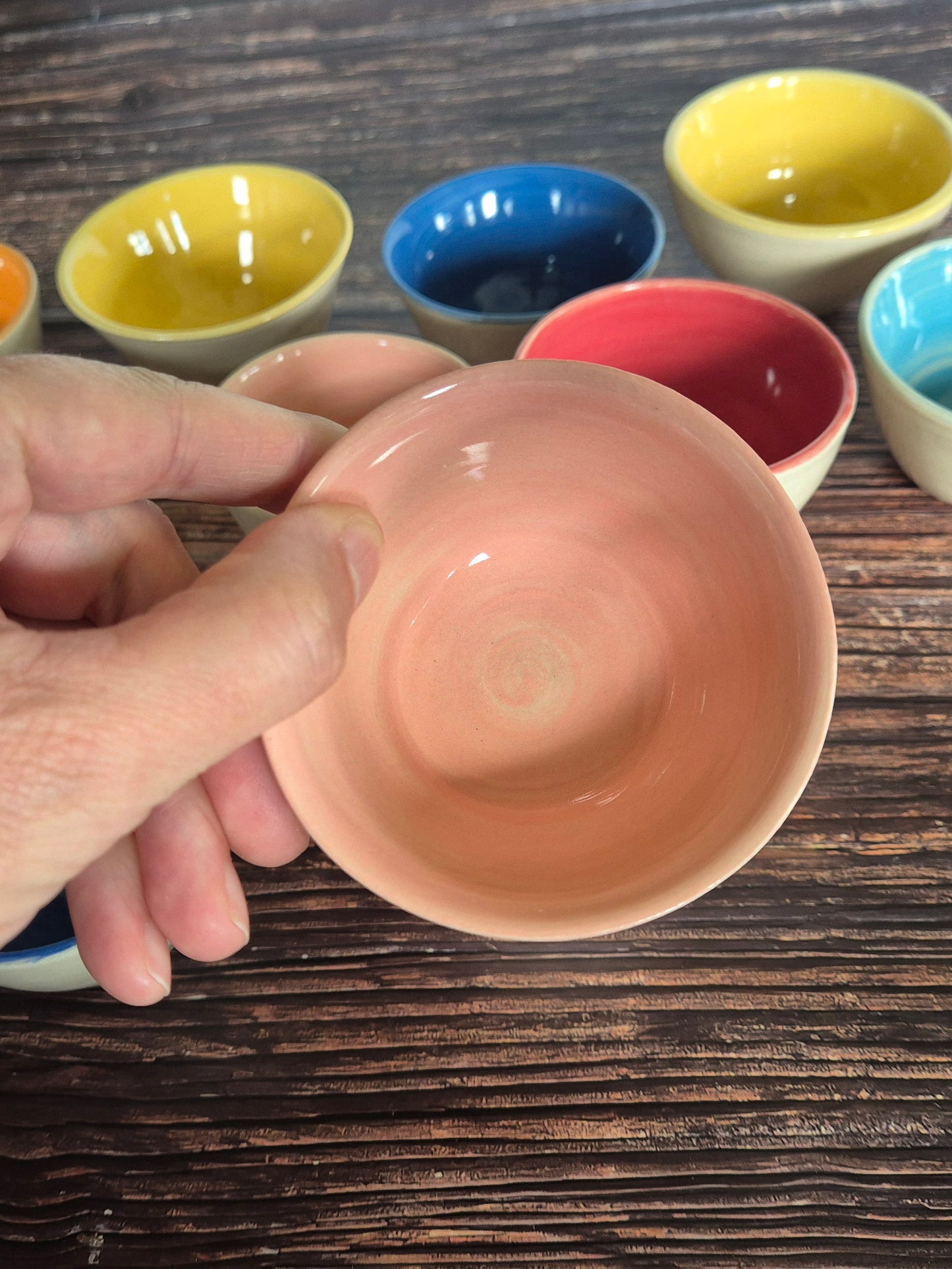 A person holding a brightly coloured condiment bowl with other colourful bowls in the background. On a wood table.