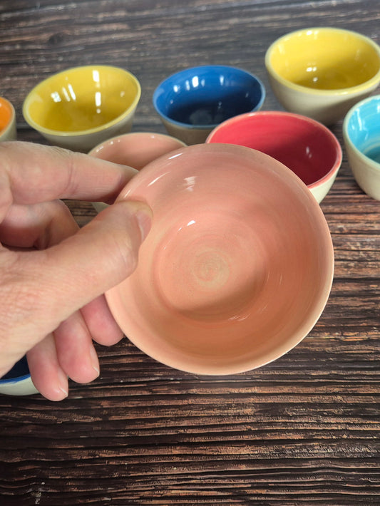 A person holding a brightly coloured condiment bowl with other colourful bowls in the background. On a wood table.
