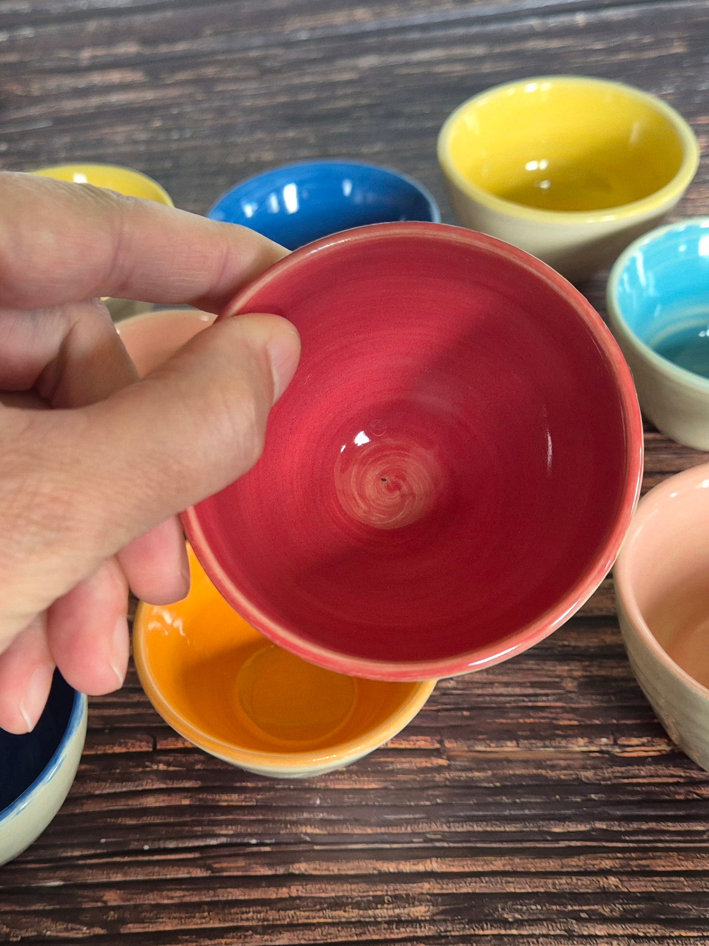 A person holding a brightly coloured condiment bowl with other colourful bowls in the background. On a wood table.