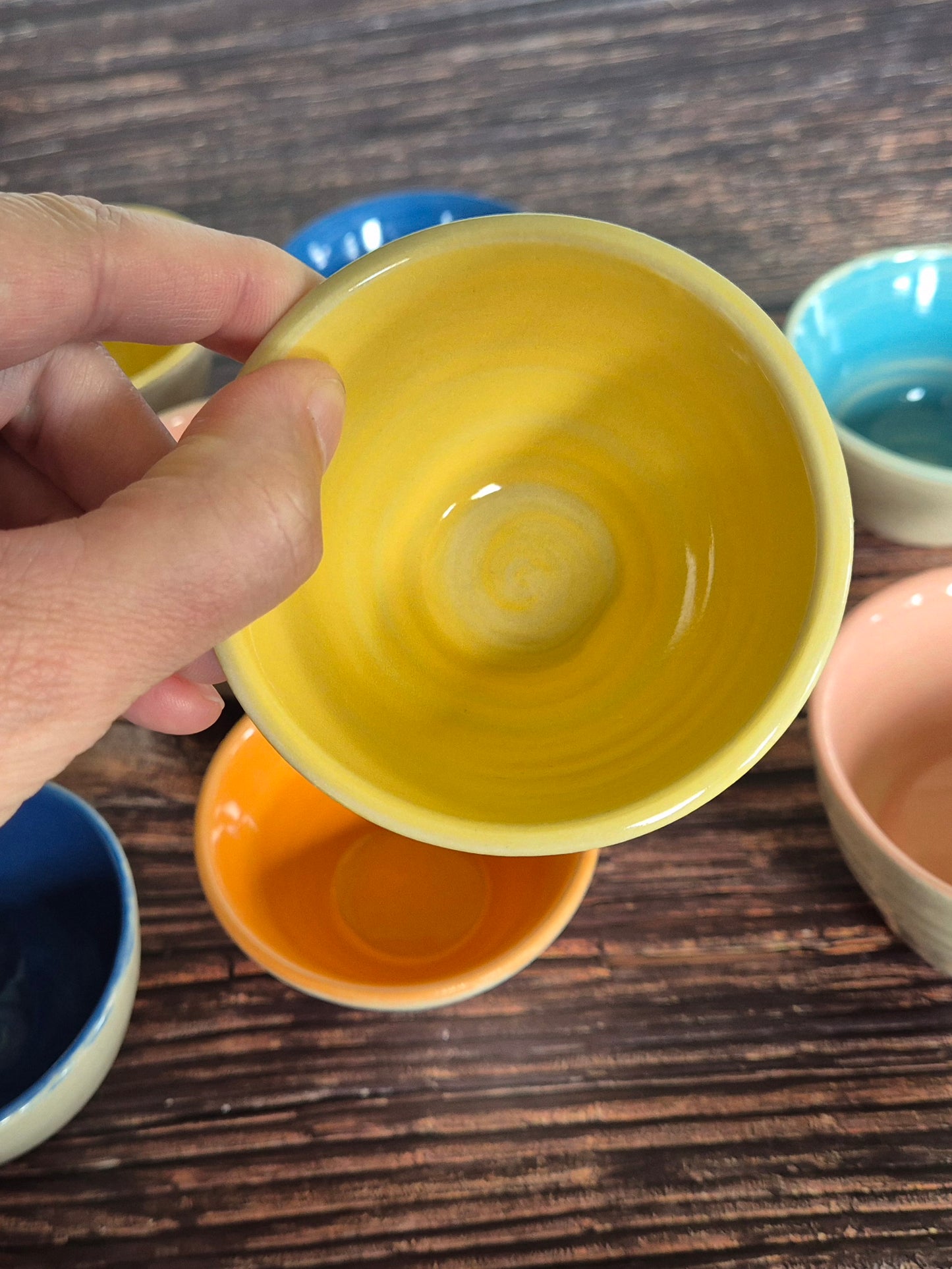 A person holding a brightly coloured condiment bowl with other colourful bowls in the background. On a wood table.