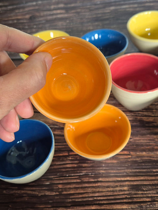 A person holding a brightly coloured condiment bowl with other colourful bowls in the background. On a wood table.
