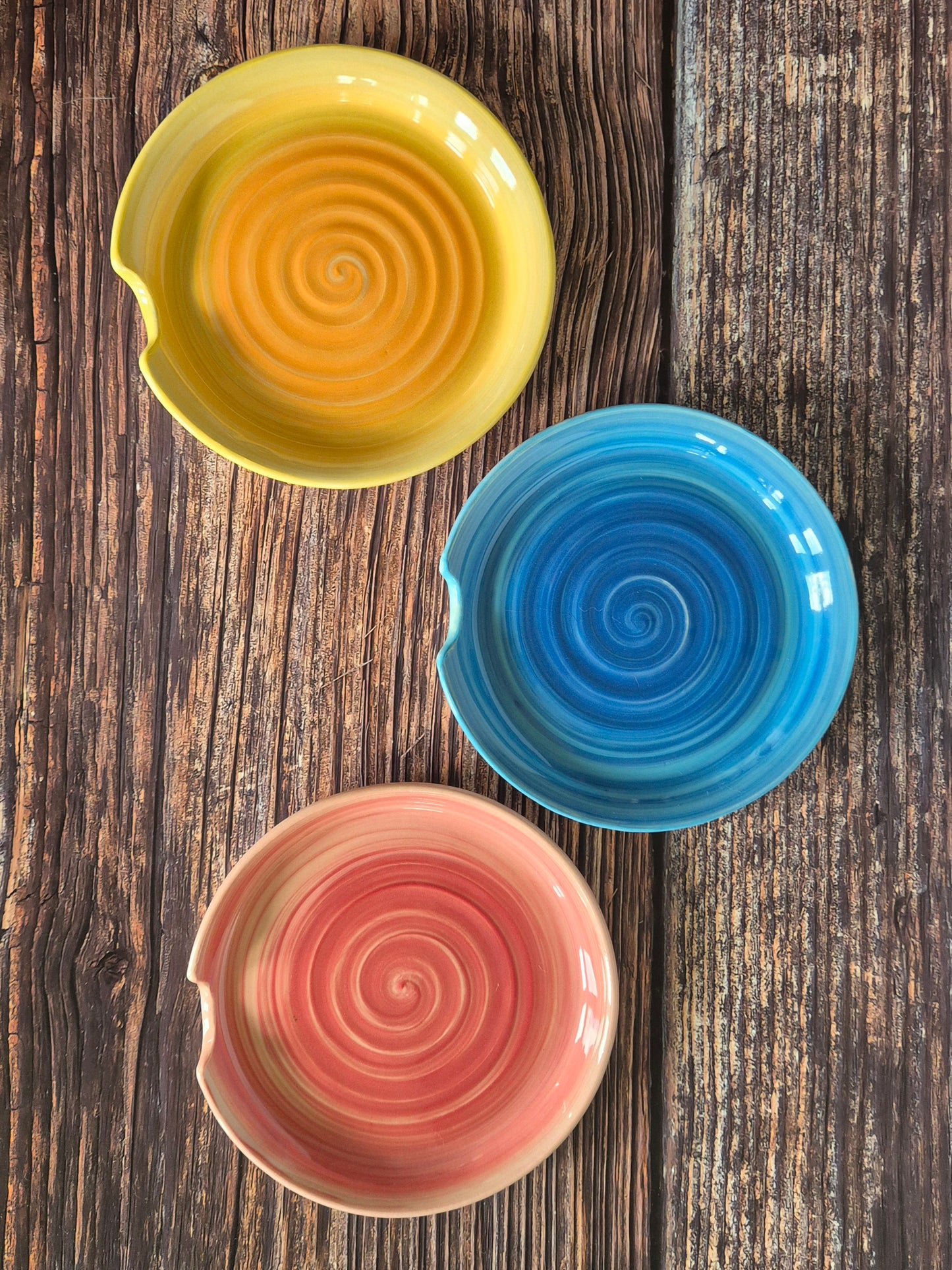 Three ceramic spoon-rests in yellow, blue, and pink with spiral patterns on a wooden surface.
