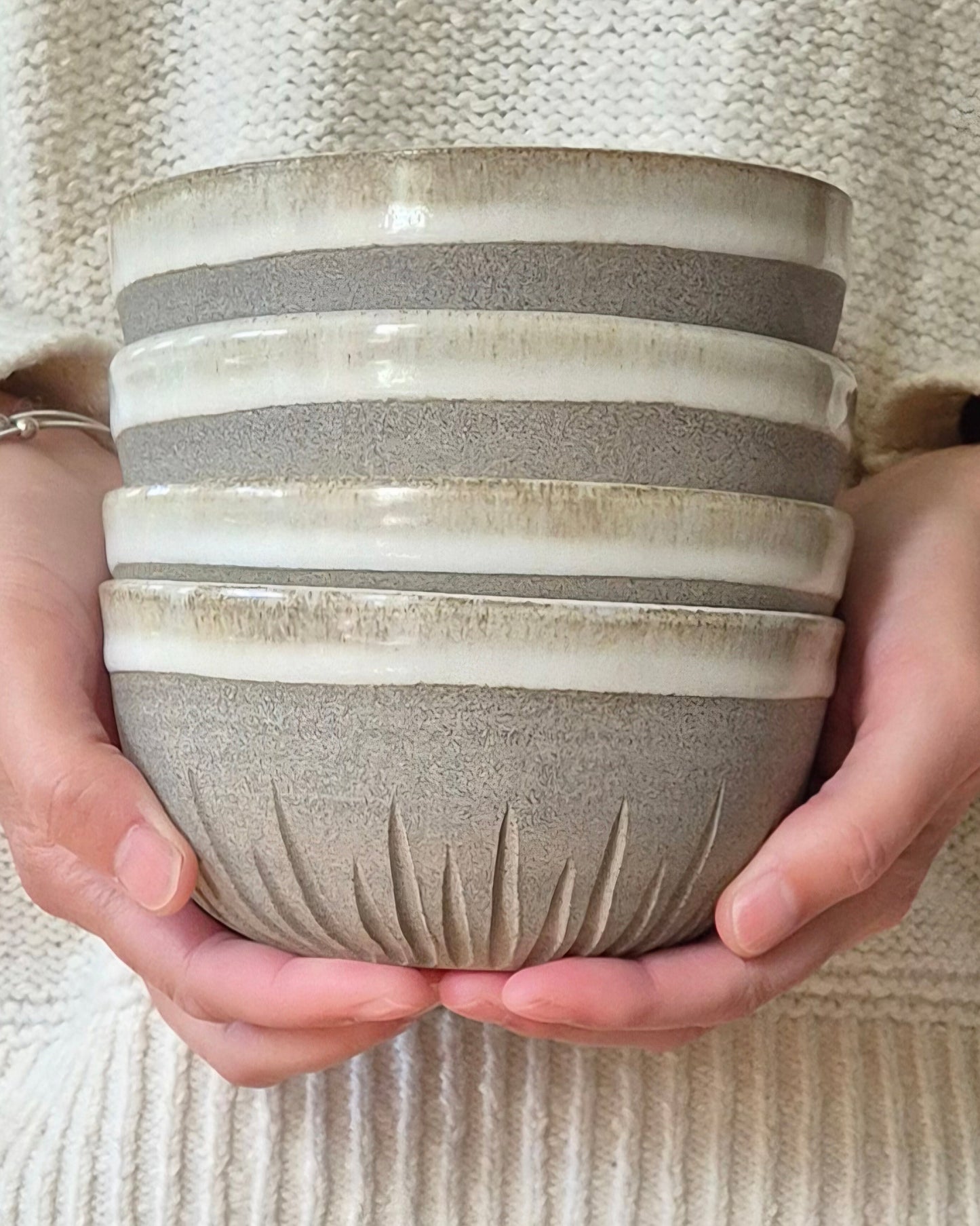 A stack of five ceramic bowls with a textured matte finish and a ribbed glazed rim, held in both hands against a person's cream-colored knit sweater.