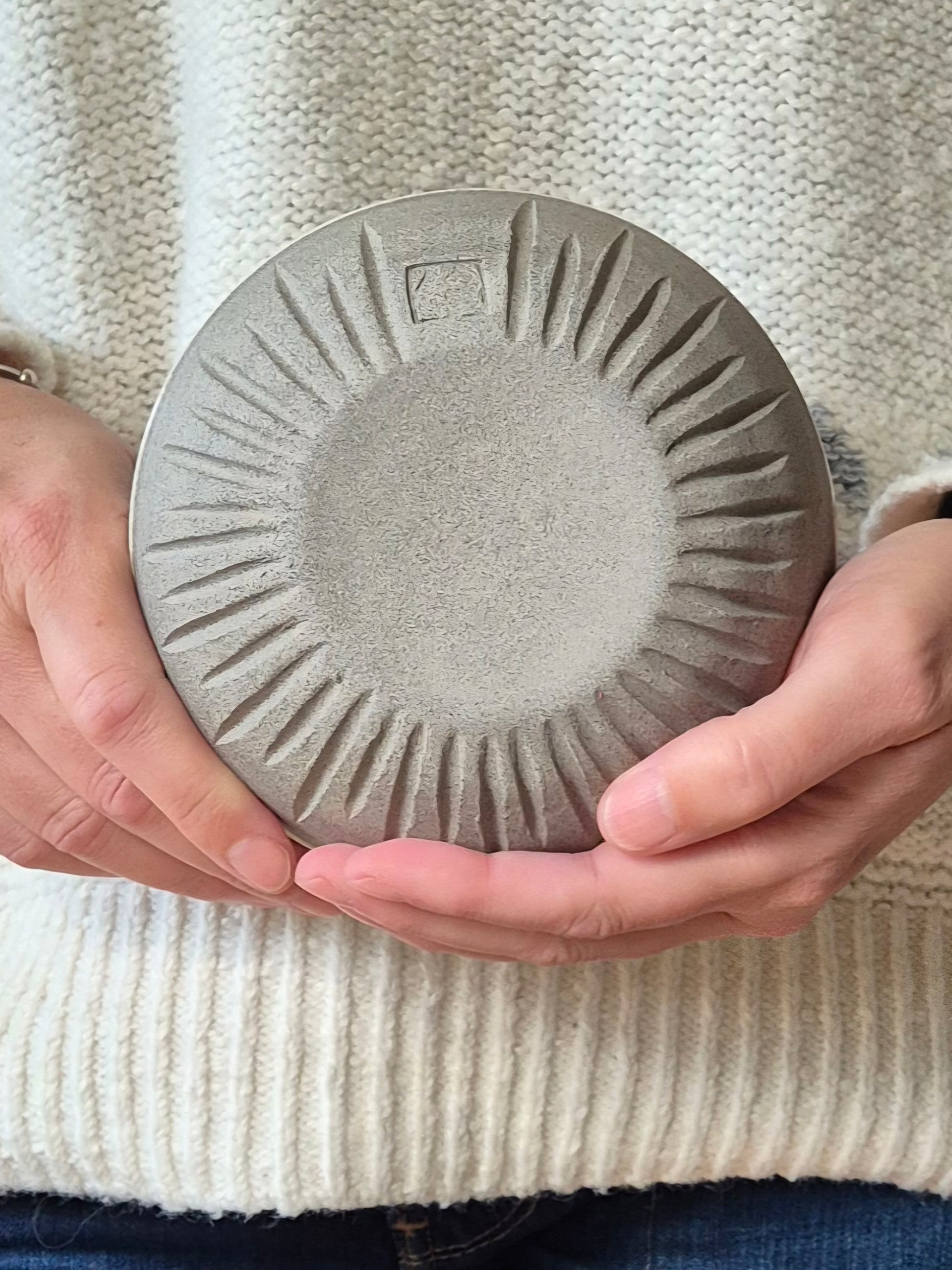 A person in a cream-colored knit sweater holds a round, textured gray ceramic plate or dish with a sunburst pattern radiating from the center.
