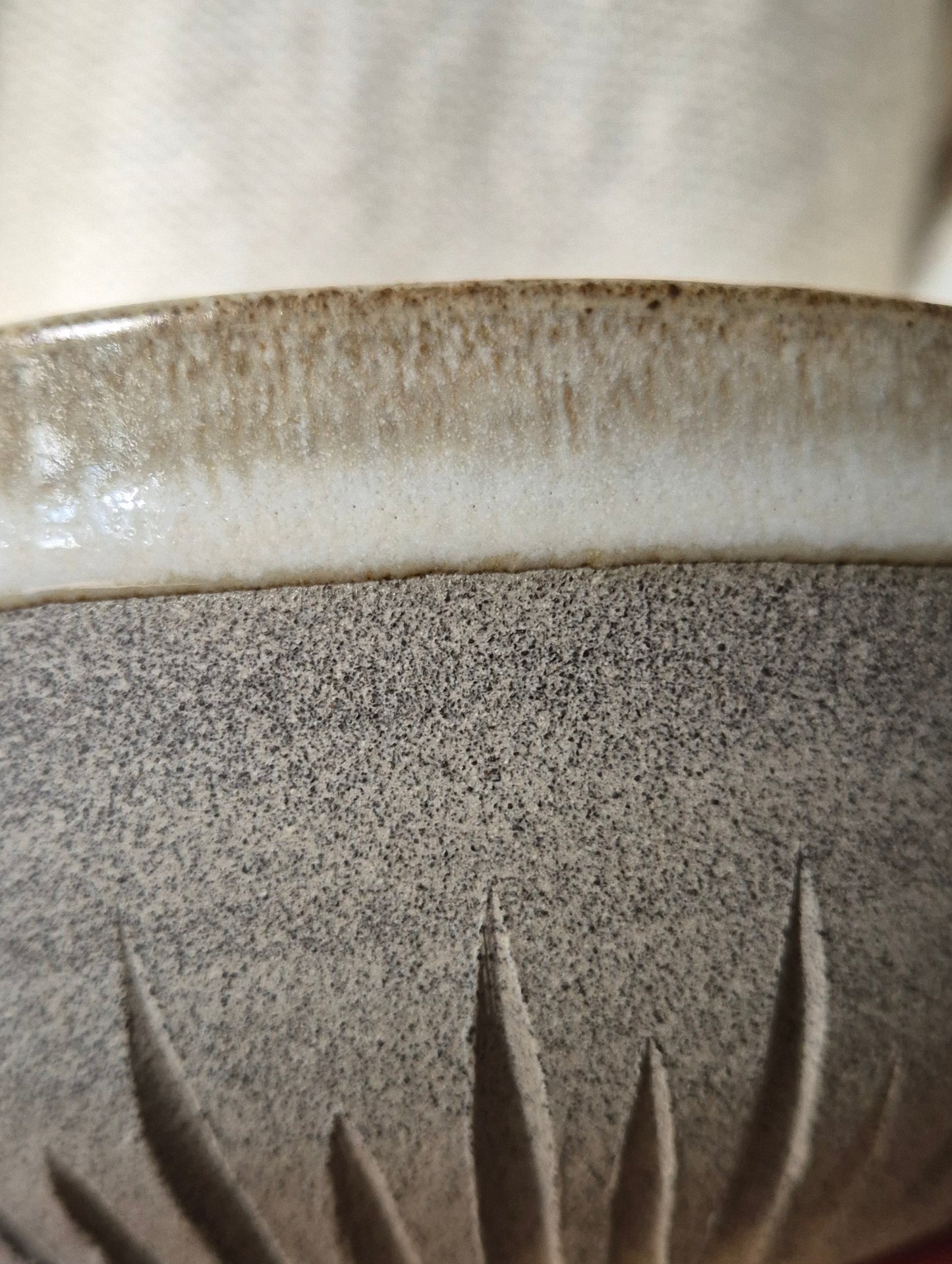 Close-up of a ceramic bowl showing a speckled gray-brown glaze, a glossy cream band around the rim, and a carved leaf pattern near the base.