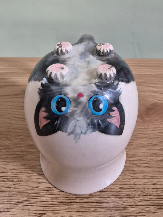 A ceramic hand-painted cat mug with a glossy finish, featuring black and white fur, large blue eyes, pink inner ears, and small pink paw pads on top, displayed on a wooden surface.