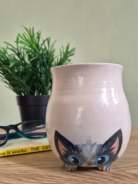A ceramic hand-painted cat-shaped mug featuring a black-and-gray cat face with blue eyes, shown on a wooden surface with a plant and glasses in the background.