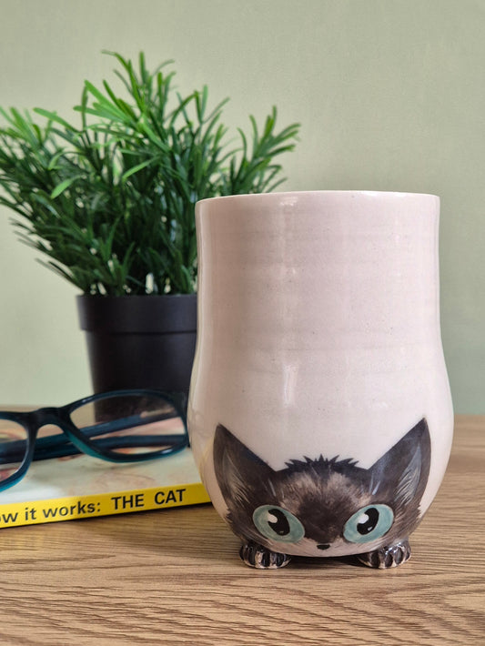 A ceramic cat-shaped cup with a glossy off-white body and a detailed illustration of a black cat peeking out from the bottom, featuring large light blue-green eyes. The cup is styled on a wooden tabletop next to a pair of eyeglasses and a potted plant against a soft green wall.