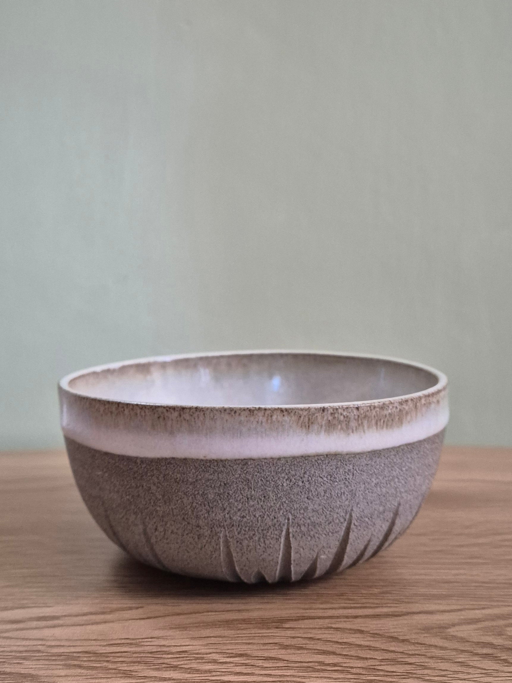 A handmade ceramic bowl with a textured speckled gray-brown exterior and a smooth, matte light beige interior, sitting on a wooden surface against a neutral gray wall.