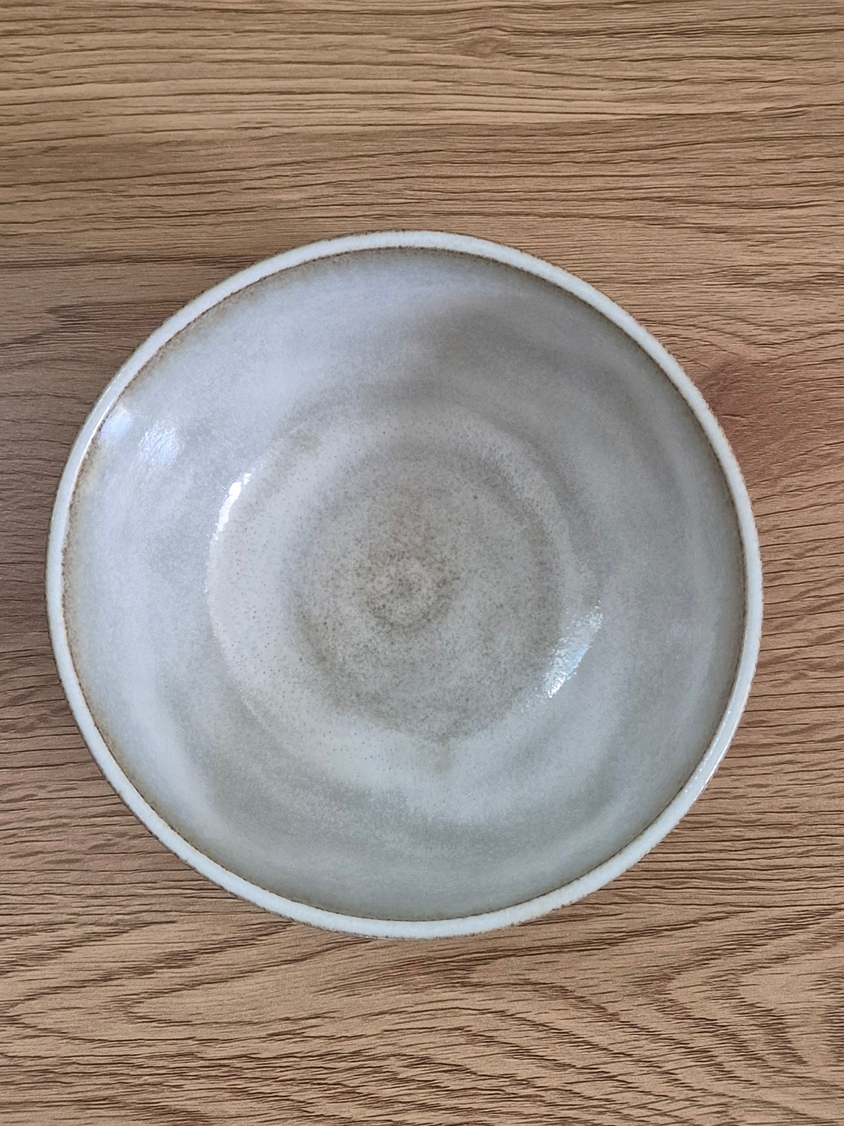 A round ceramic bowl with a matte, speckled light gray and off-white glaze, photographed from directly above on a light brown wooden surface.
