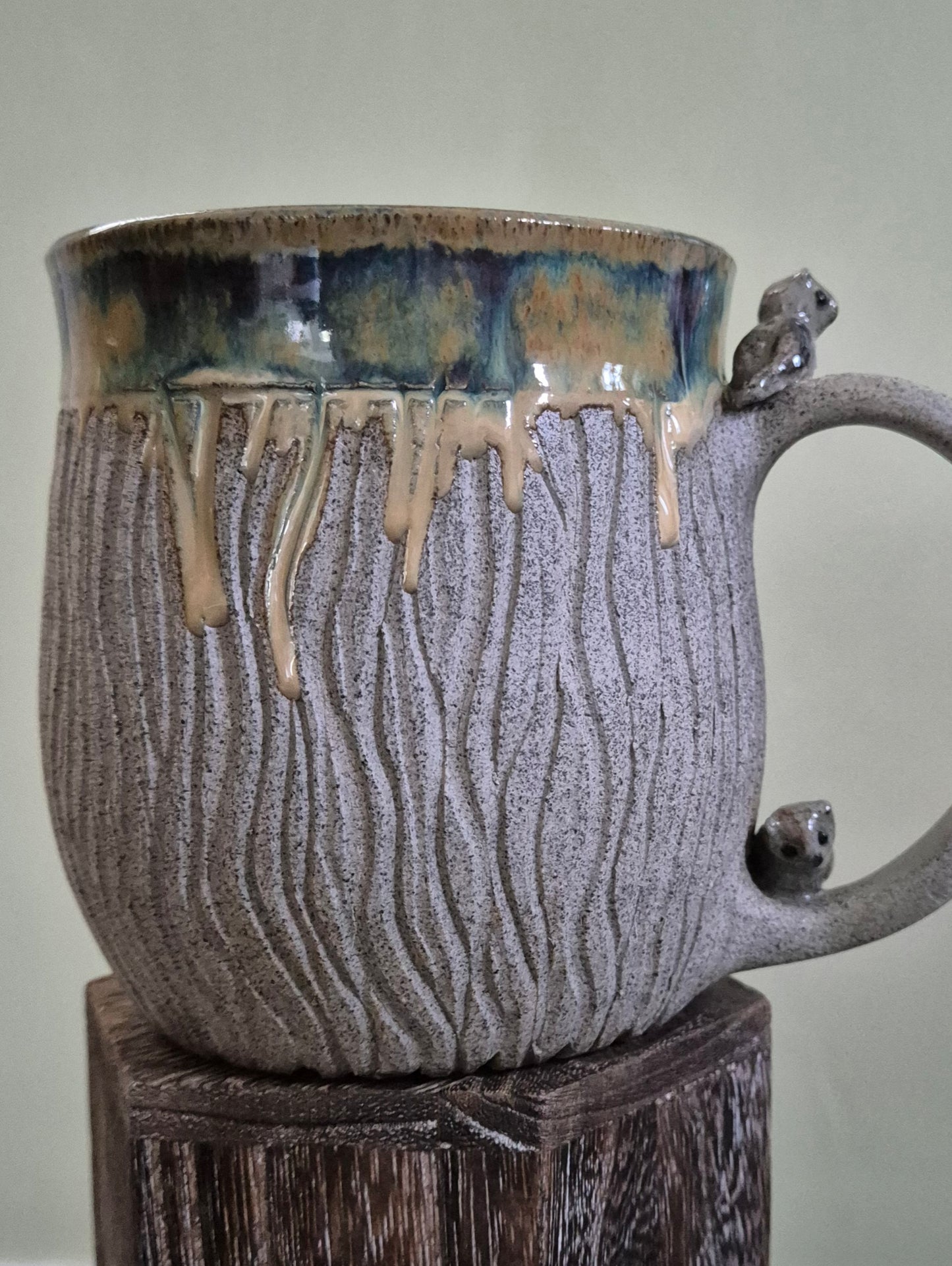 A handmade ceramic mug with a textured, ribbed gray body, a dripping blue and beige glaze at the rim, and small sculpted owl figures on the handle, displayed on a rustic wooden block against a neutral background.