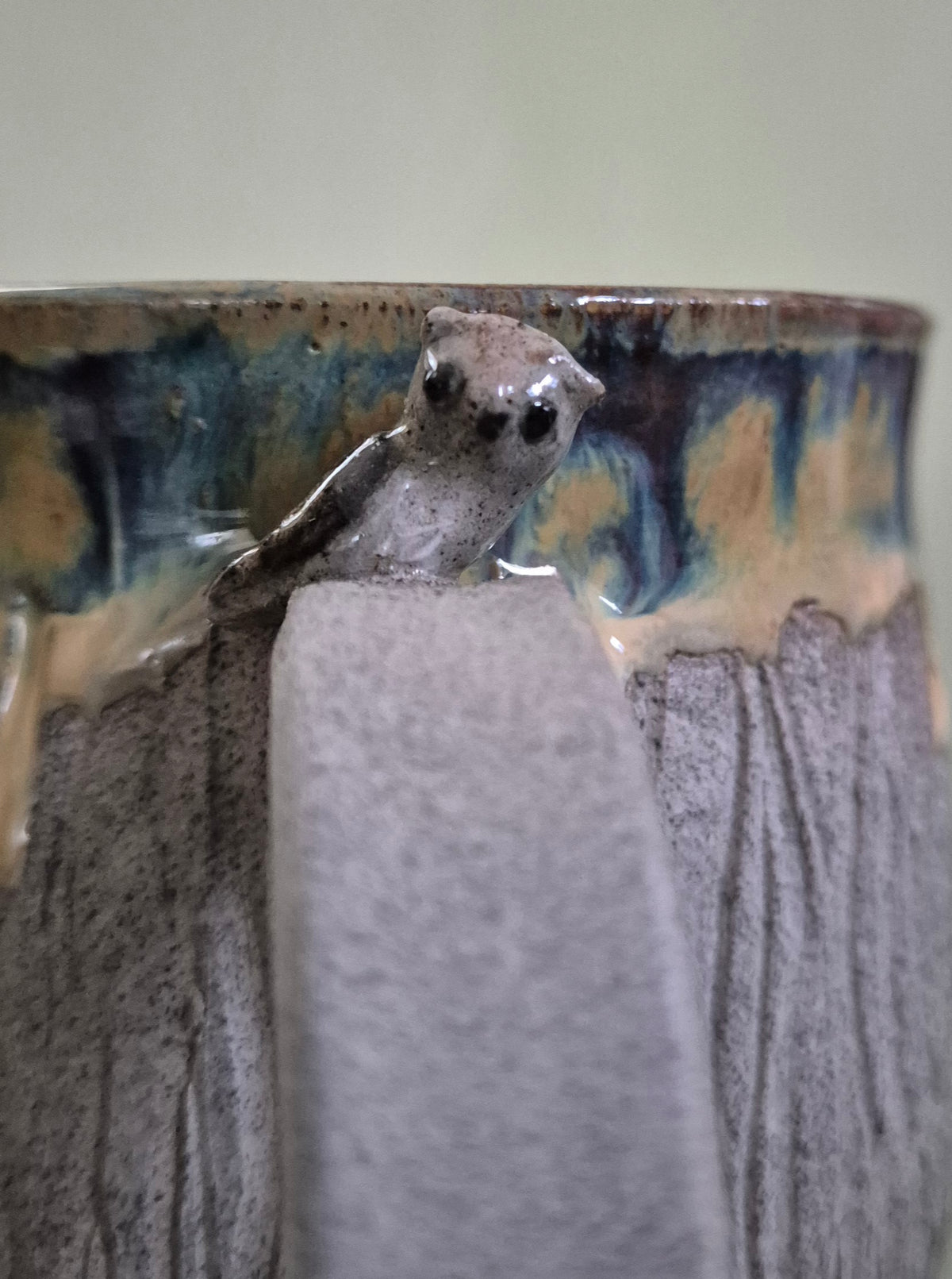 A close-up of a ceramic mug featuring a sculpted owl figure attached to the side, with a textured gray body and a glazed rim showing blue, brown, and beige drips.