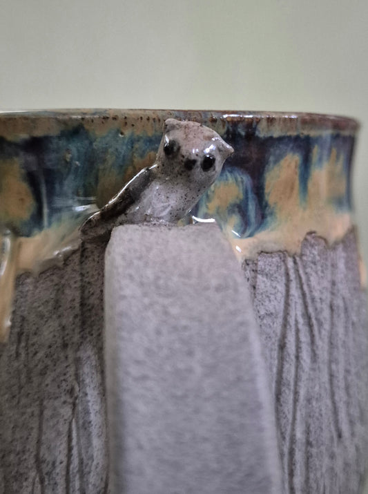 A close-up of a ceramic mug featuring a sculpted owl figure attached to the side, with a textured gray body and a glazed rim showing blue, brown, and beige drips.