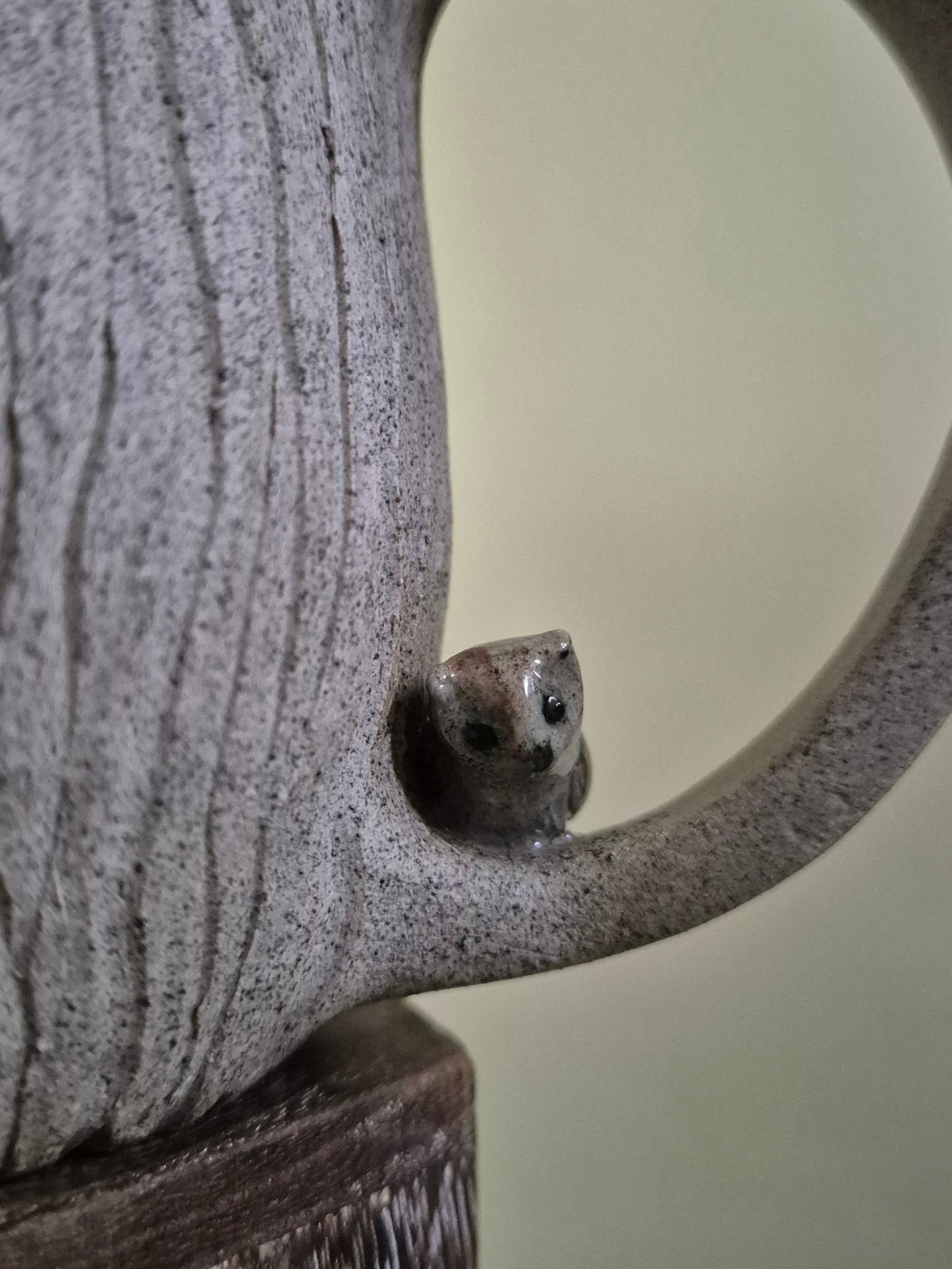 Close-up of a speckled gray ceramic pitcher with a small owl figurine perched on the handle, set against a soft beige background.