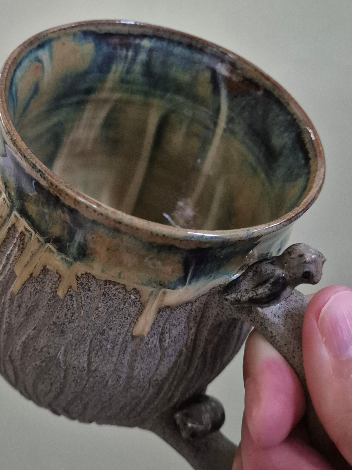 A close-up of a handmade ceramic mug with a textured bark-like handle and a marbled glaze, held by a hand against a neutral background.