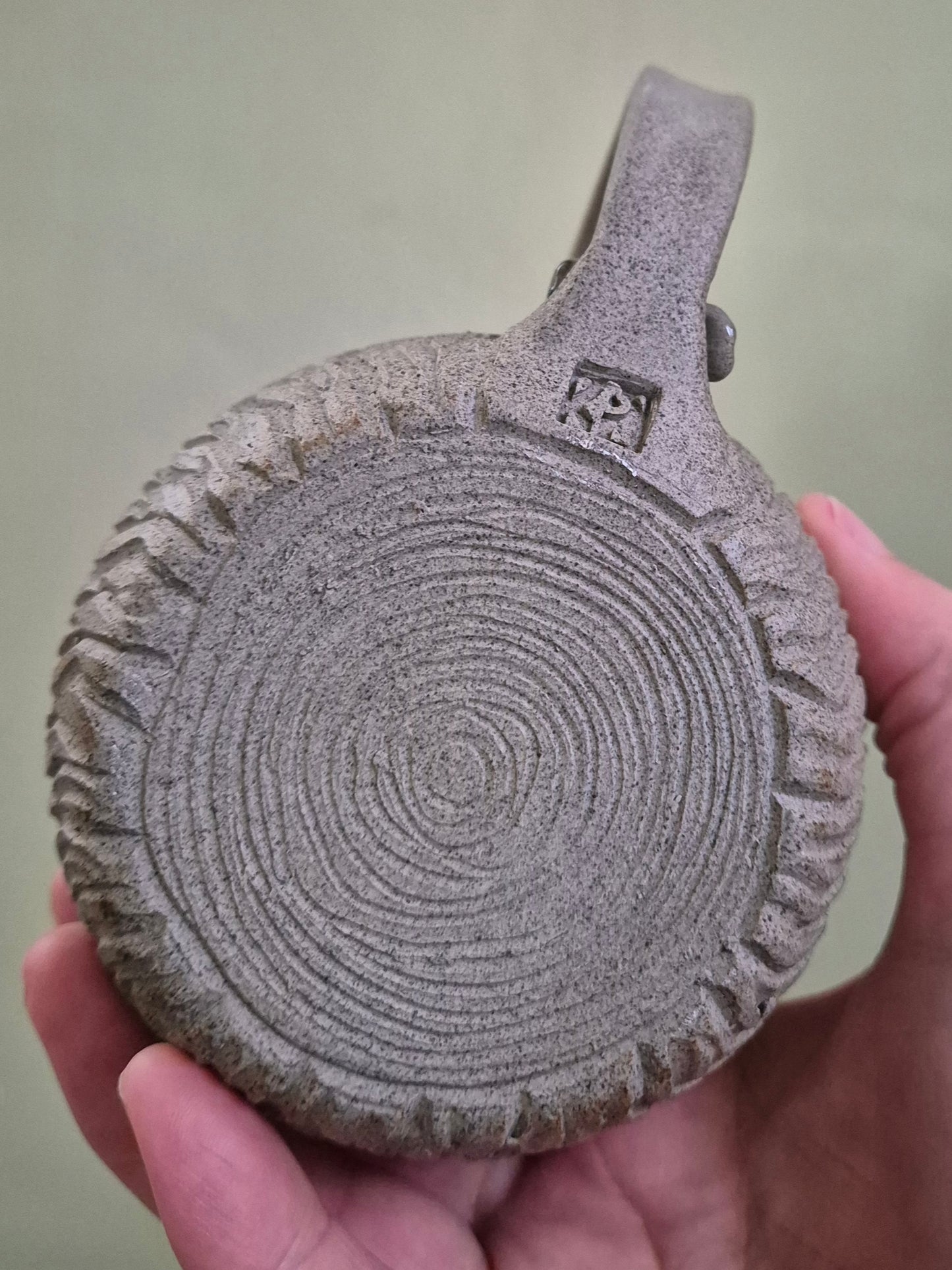 A hand holding a round, textured gray ceramic mug with a loop handle, featuring a carved concentric circle pattern on the base and a small carved logo in the upper right.
