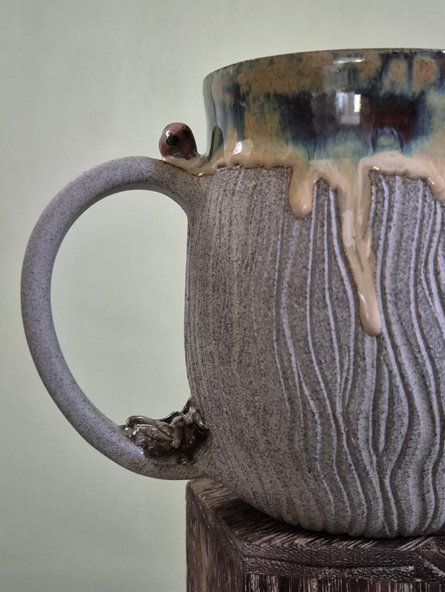 A close-up of a handmade ceramic mug with a textured, ribbed gray body, a dripping beige glaze, and a dark brown glazed rim, resting on a wooden block against a neutral background.