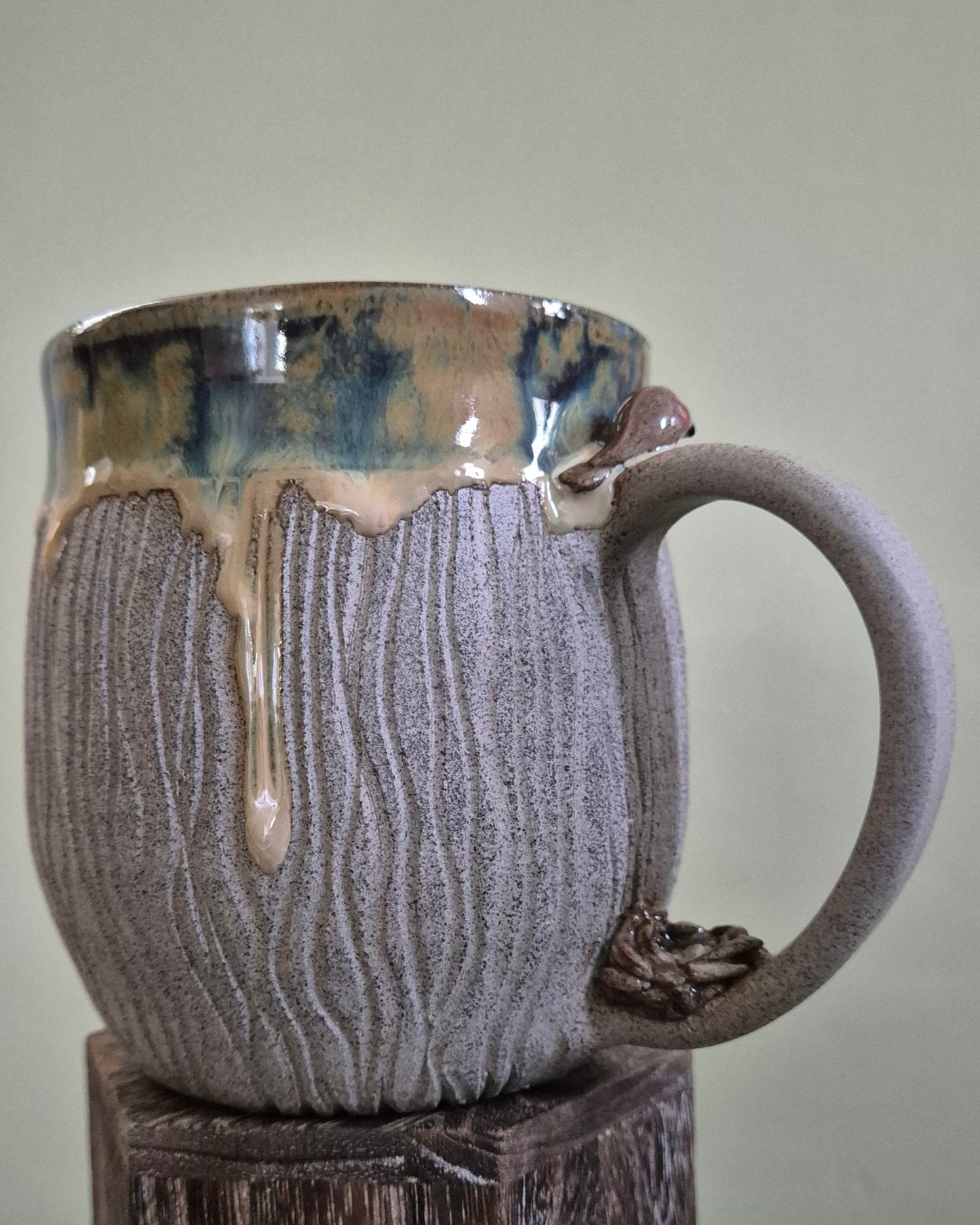 A handmade ceramic mug with a textured gray body, a dripping creamy gold and blue-green glazed rim, and a dark brown handle, displayed on a rustic wooden pedestal against a light green wall.