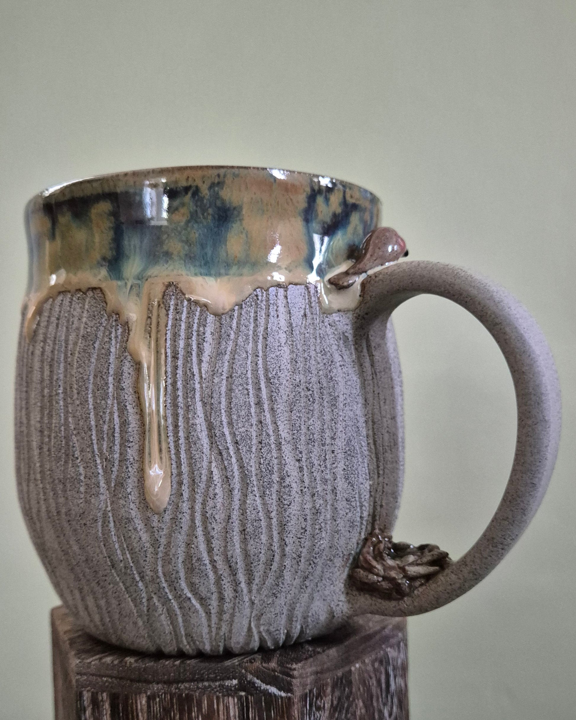 A handmade ceramic mug with a textured gray body, a dripping creamy gold and blue-green glazed rim, and a dark brown handle, displayed on a rustic wooden pedestal against a light green wall.