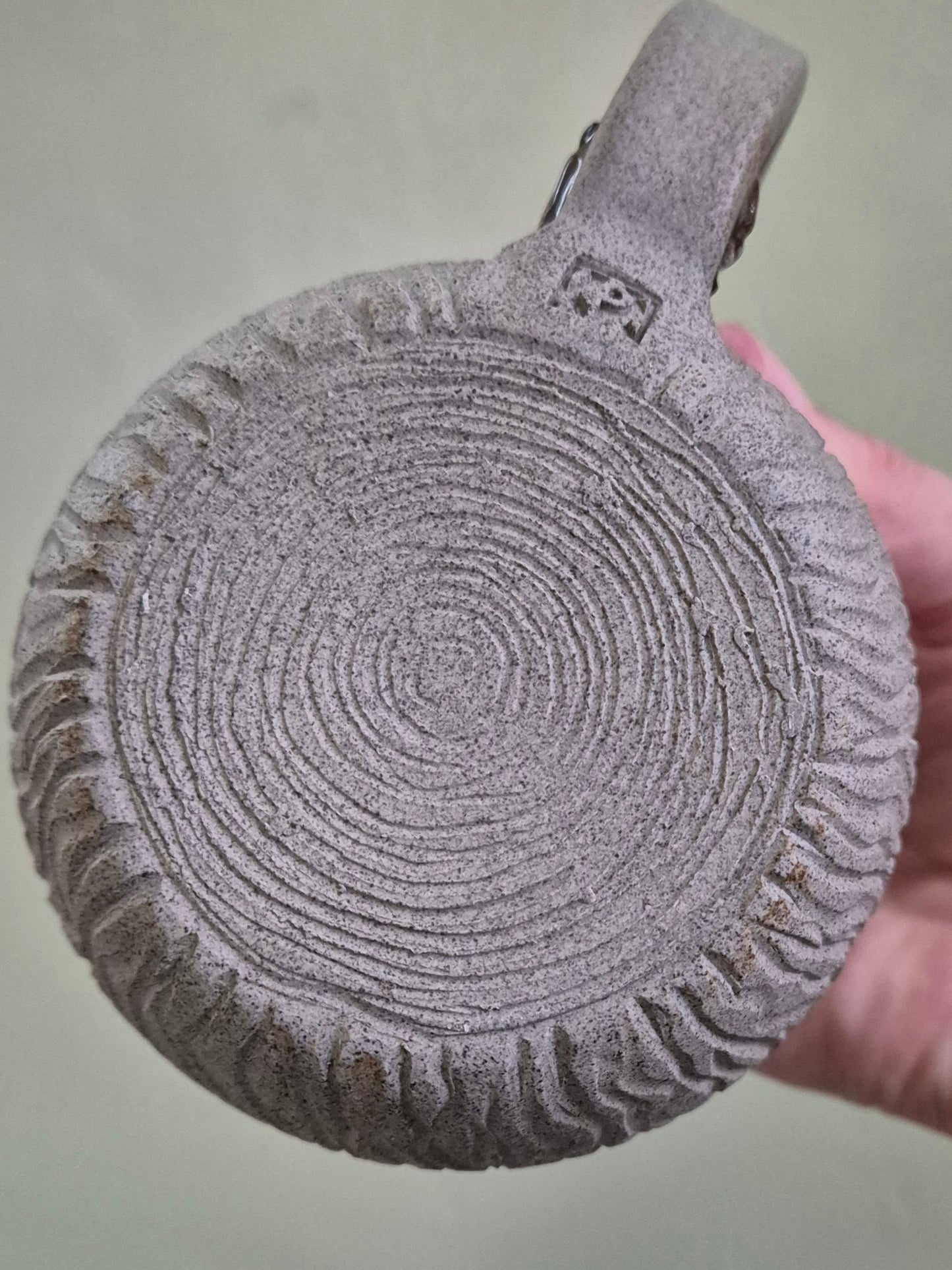 Base of a round, textured gray ceramic mug with concentric circular grooves and a ridged outer edge, held by a hand against a muted green background.