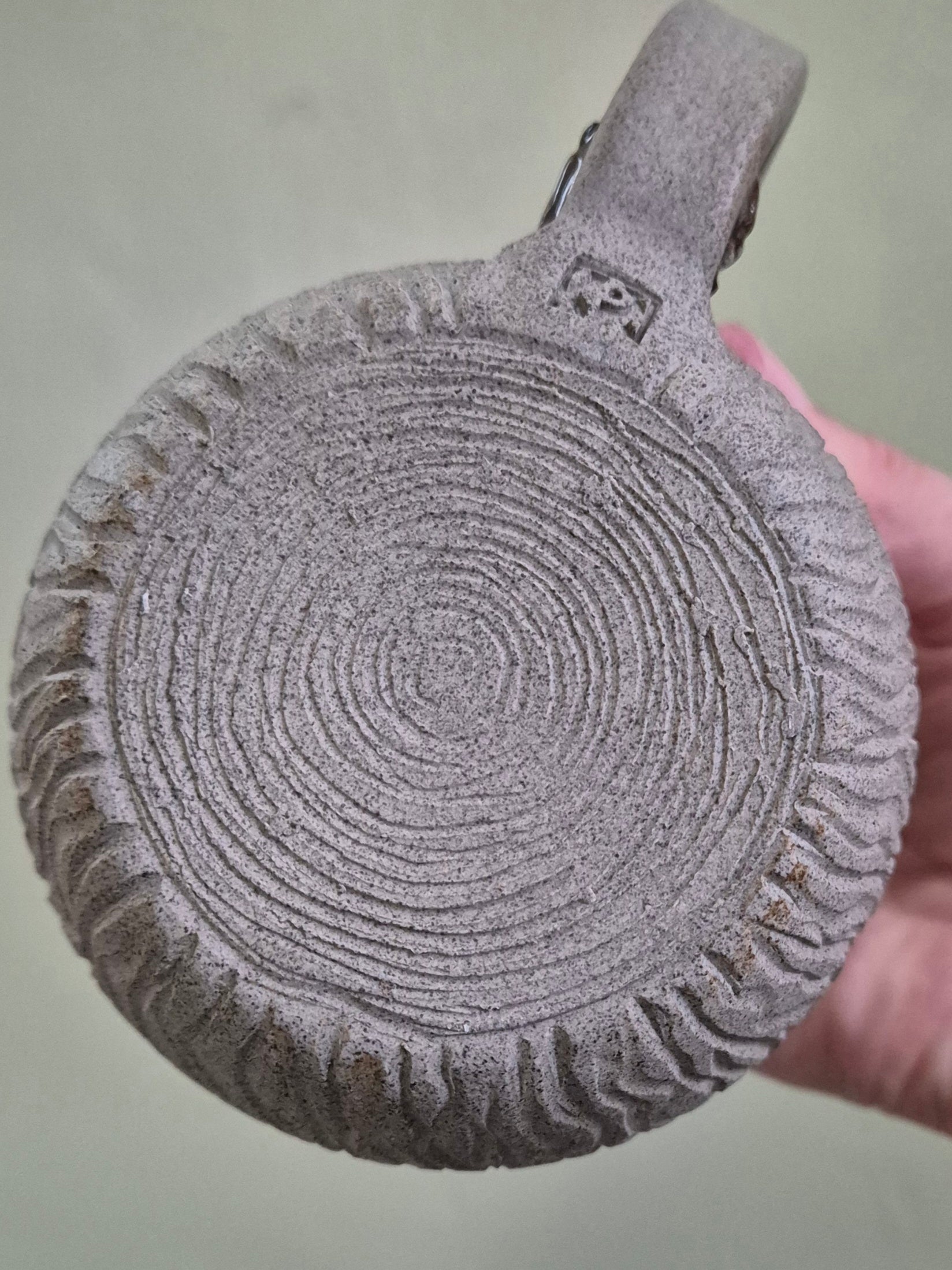 A round, textured gray stone or ceramic plate with concentric circular grooves and a ridged outer edge, held by a hand against a muted green background.