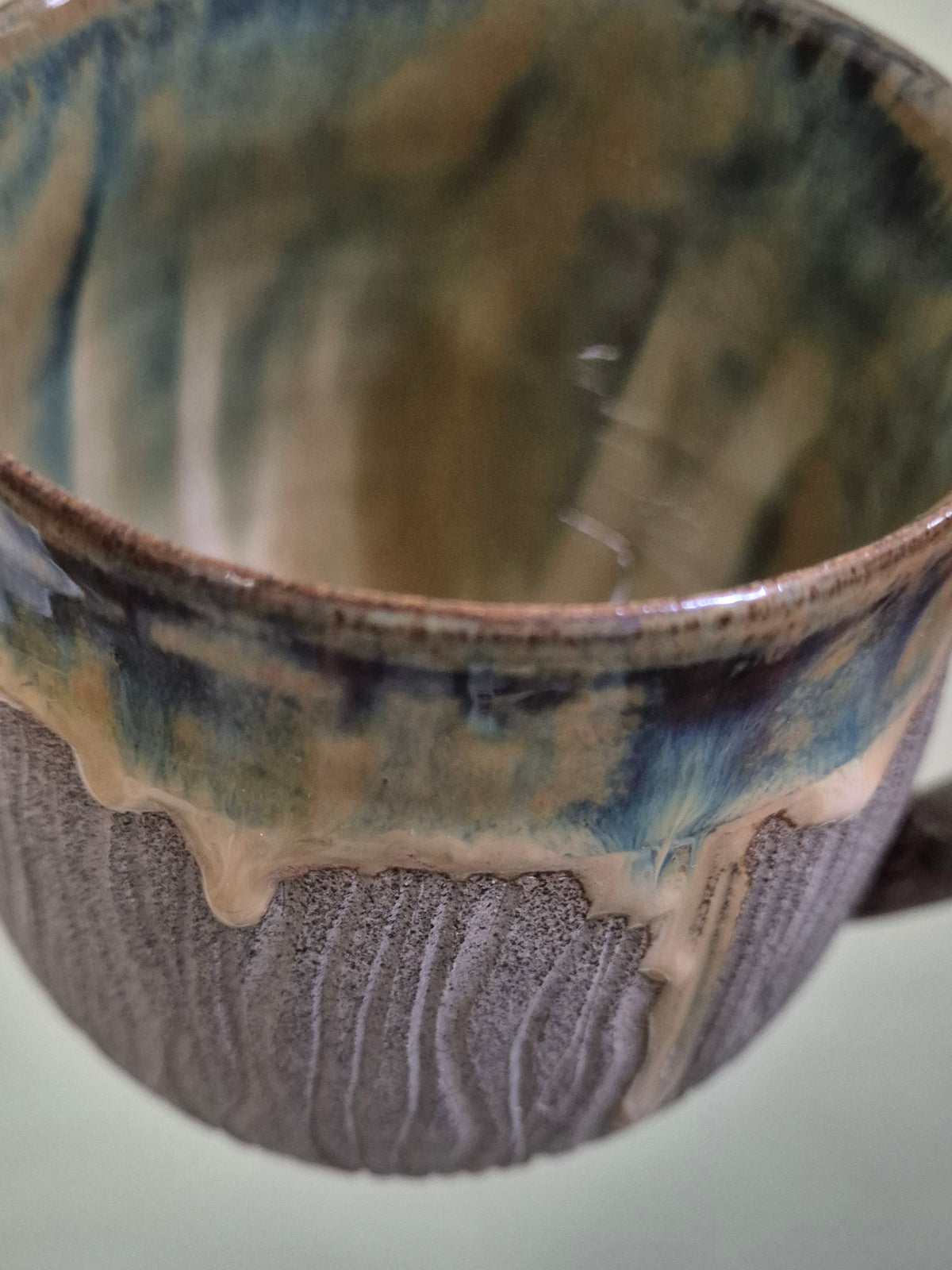 Close-up of a handmade ceramic mug with a textured, ribbed exterior in a grayish-purple glaze and a blue, brown, and cream drip-glaze interior.
