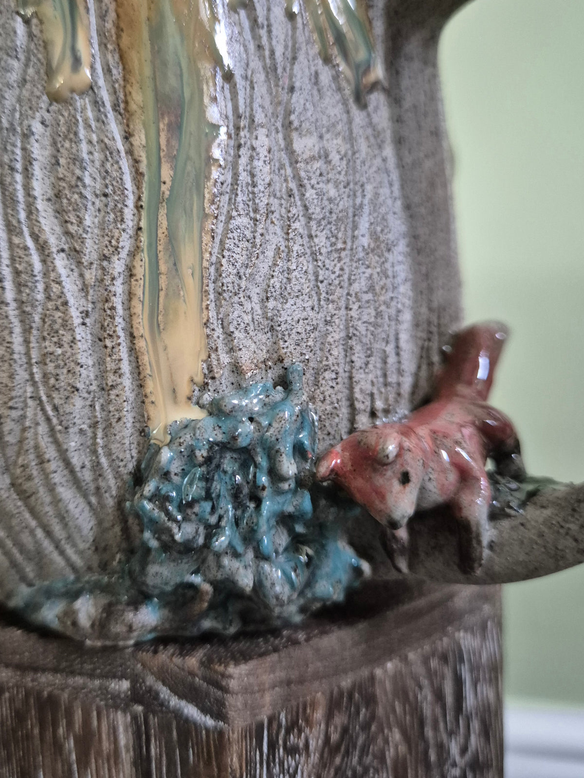 Close-up of a sculptural ceramic piece featuring a textured gray and gold vertical form, a glossy teal blue abstract cluster, and a small reddish-brown fox figurine on a branch-like extension, mounted on a dark wooden base.