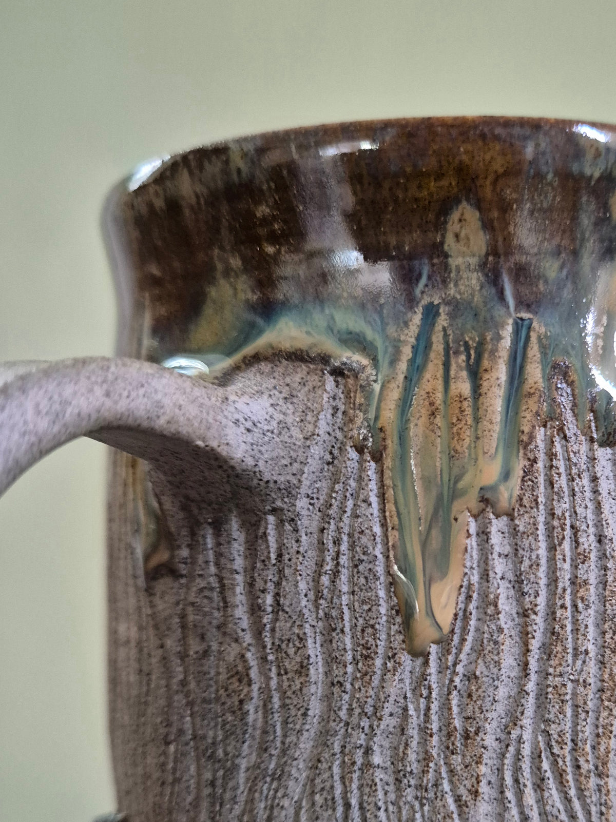 Close-up of a handmade ceramic mug with a textured, bark-like surface and dripping glaze in earthy brown, beige, and green tones.