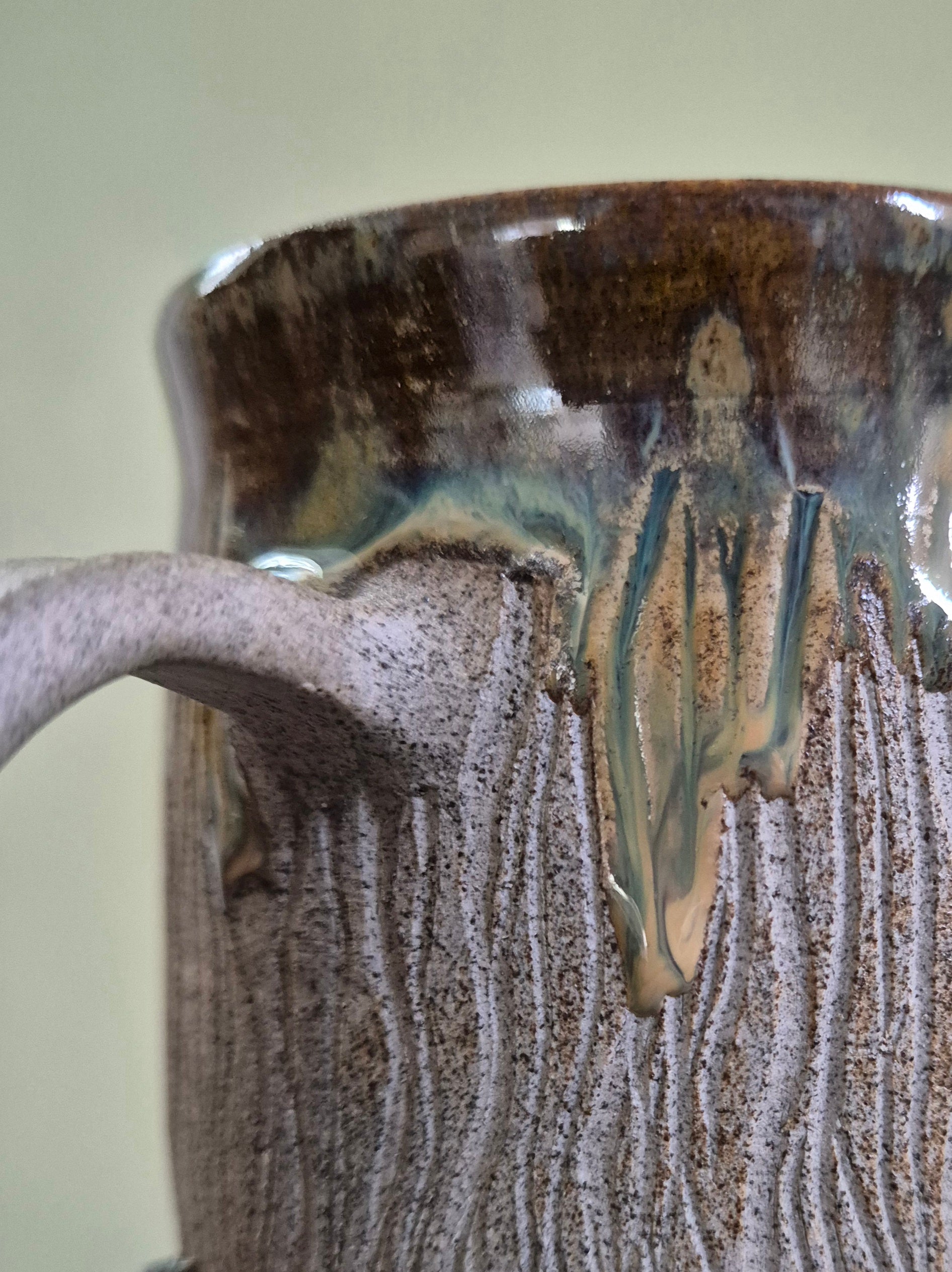 Close-up of a handmade ceramic mug with a textured, bark-like surface and dripping glaze in earthy brown, beige, and green tones.