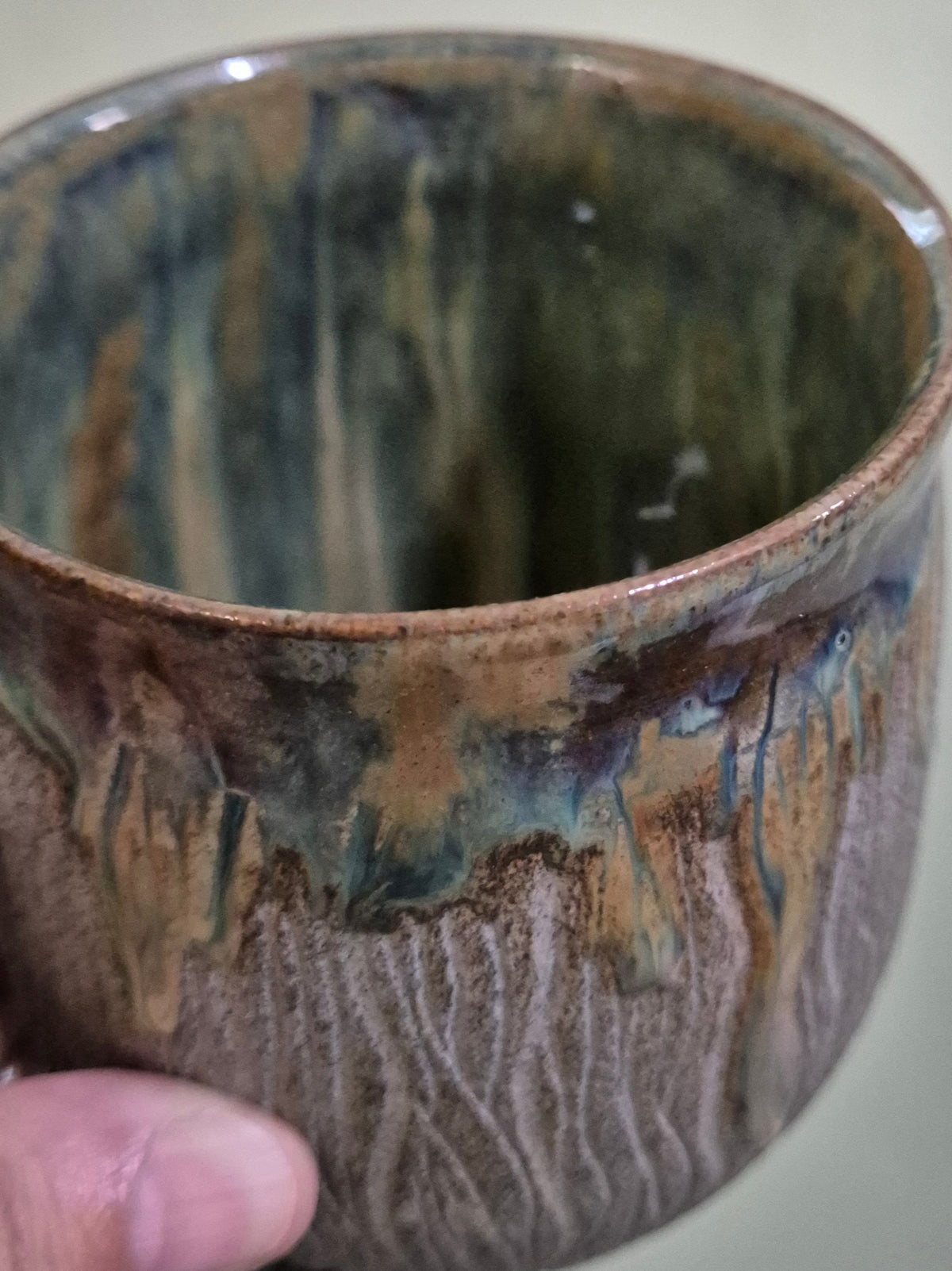 A close-up of a handmade ceramic cup held by a hand, showcasing a rustic, dripping glaze with earthy brown, green, and blue tones.
