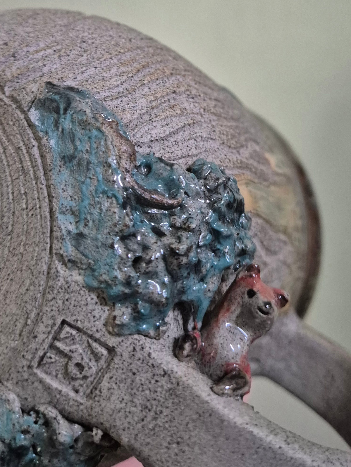 Close-up of a textured ceramic sculpture or decorative vessel with a speckled gray base, glossy teal-blue abstract relief, and a small reddish-brown fox figure near the right side.