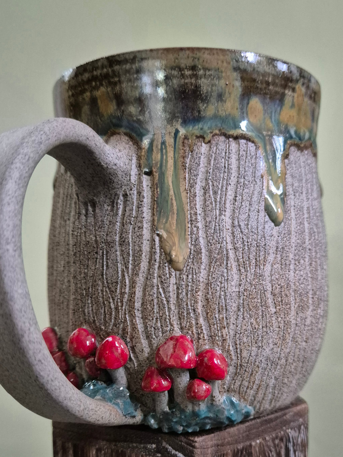 A handmade ceramic mug with a textured, bark-like surface in gray-brown tones, featuring a dripping metallic green and gold glaze at the rim and small red mushrooms growing out of the base.