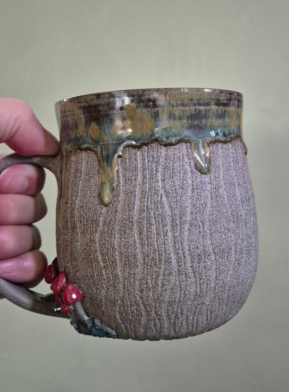 A handmade ceramic mug with a textured, bark-like surface in earthy brown and gray tones, featuring a dripping metallic gold and green glaze at the rim and small red toadstools on the handle, held by a hand against a plain beige background.