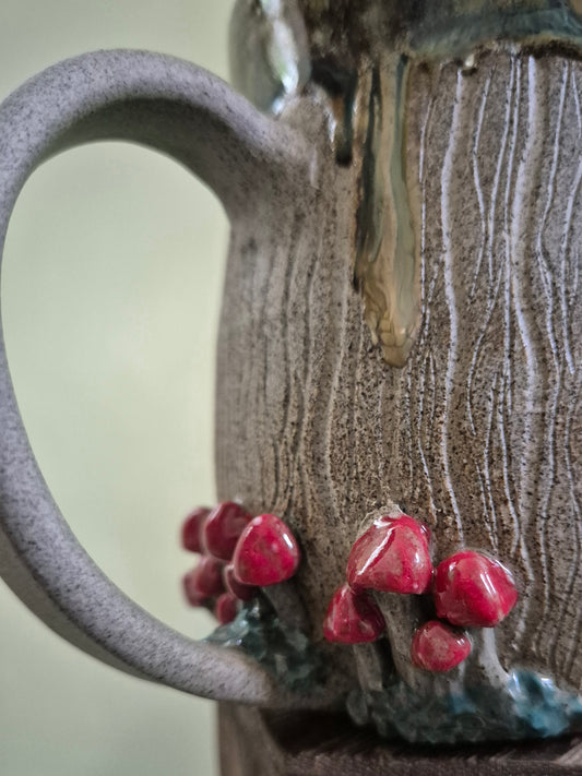 Close-up of a handmade ceramic mug with a textured, bark-like surface, a large handle, and glossy red mushroom decorations near the base.