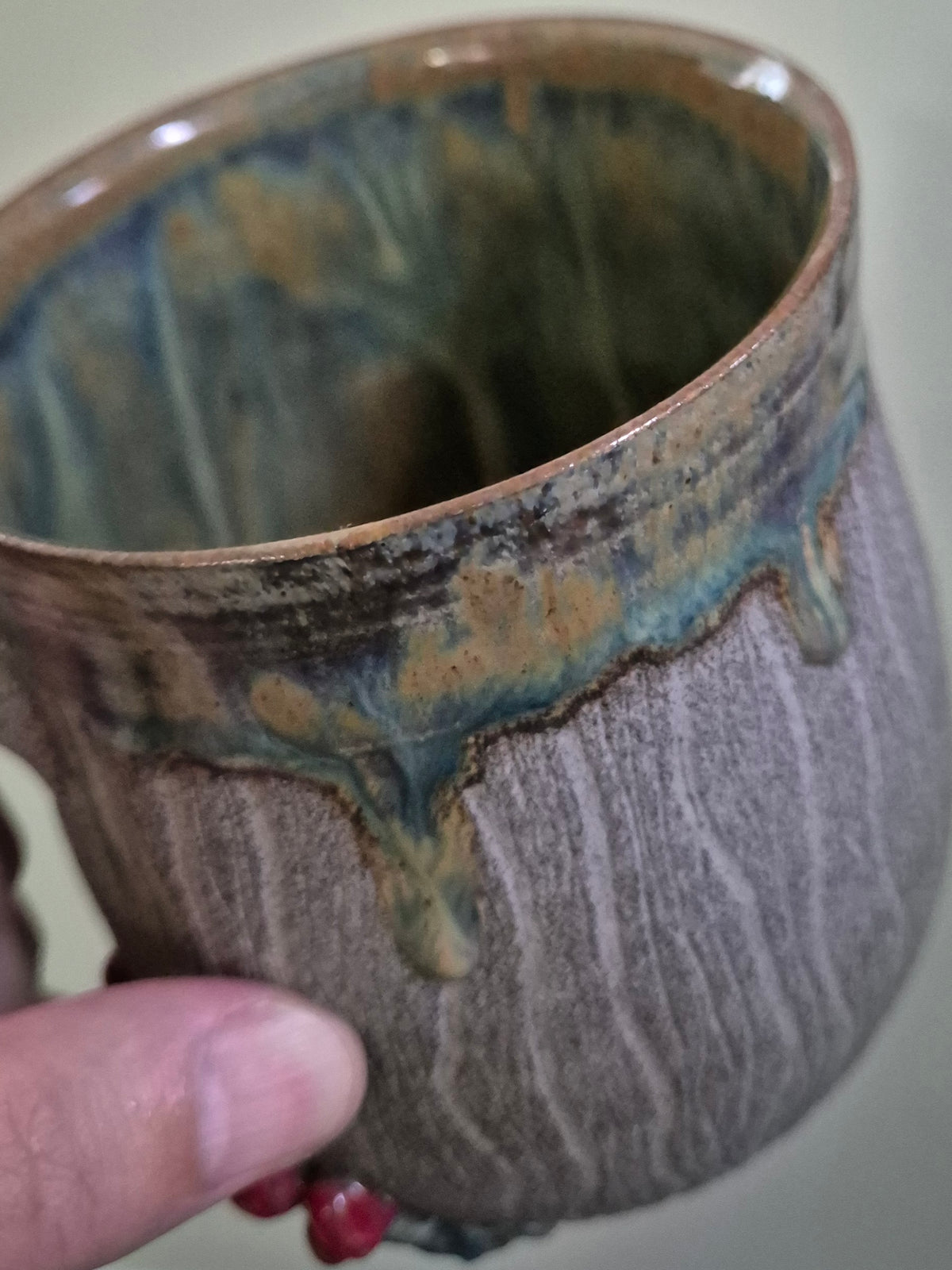 A close-up of a handmade ceramic mug with a textured, earthy brown exterior and a dripping blue-green glaze, held by a hand.