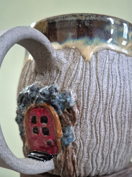 Close-up of a ceramic mug with a textured, bark-like exterior in beige and brown, featuring a small red door and blue-gray stone wall detail on the side.