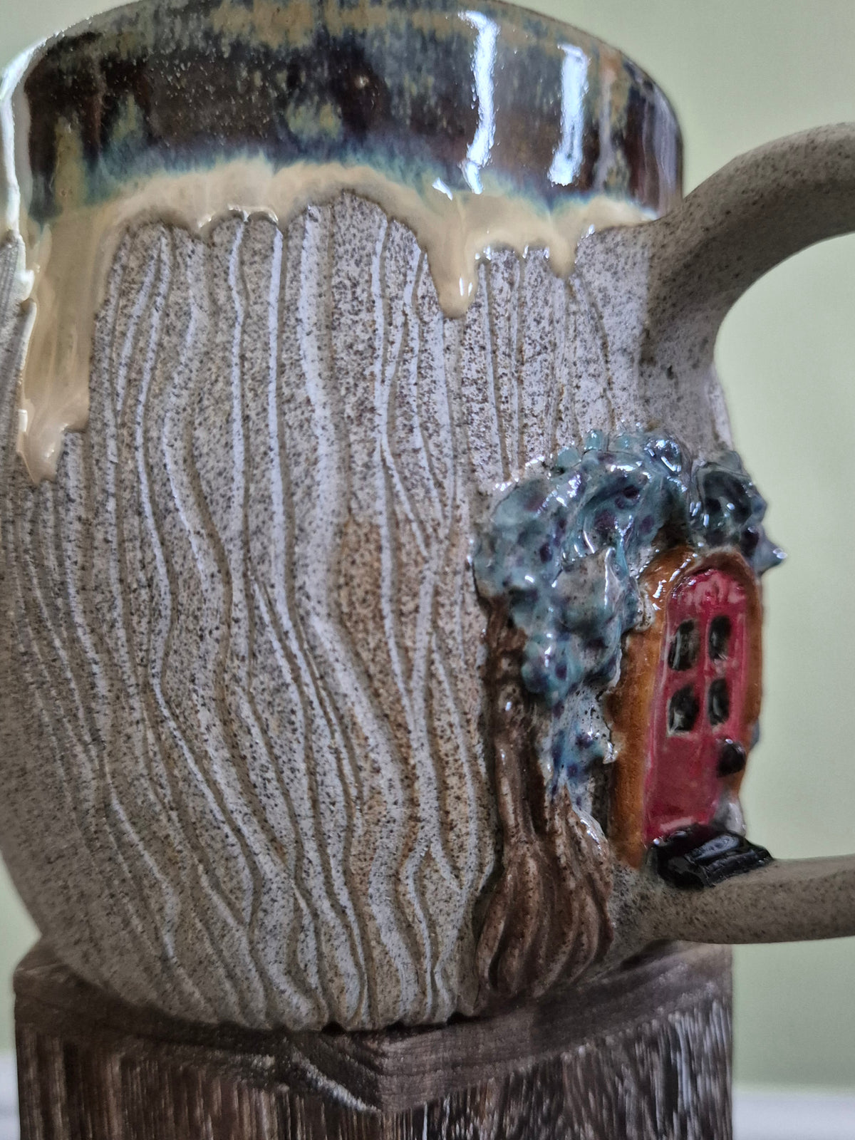 A close-up of a handmade ceramic mug with a textured, wood-grain-like glaze, featuring a small red fairy house under arched branches. Blue-gray glaze around the rim, set against a soft light green background.