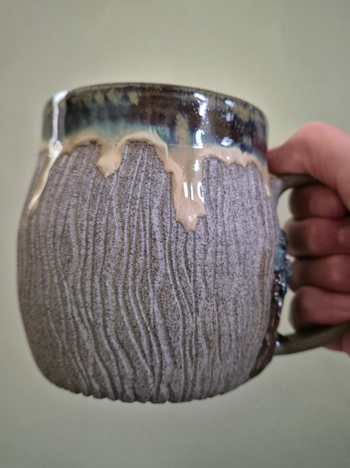 A handmade ceramic mug with a textured, ribbed gray and brown glaze, held by a hand against a plain light green background.
