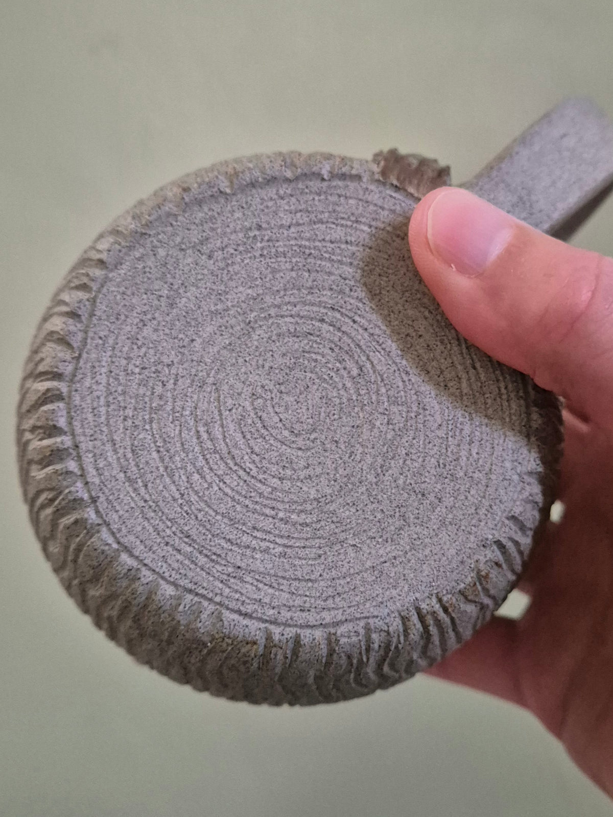 A close-up of a hand holding a round, textured gray mug with concentric circular patterns and a ribbed edge, against a plain light background.